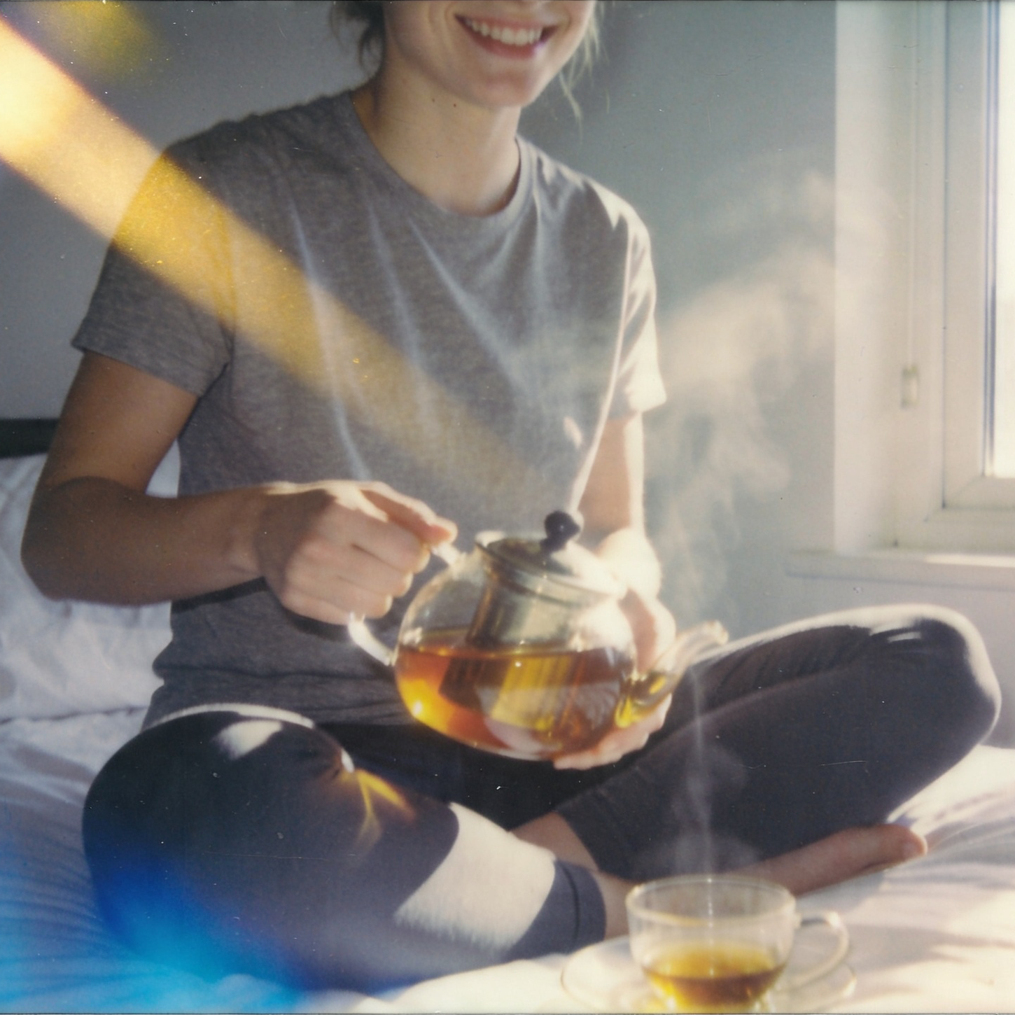 Person sitting on a bed holding a teapot and a cup of tea, with a blurred background.